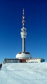A large telecommunications tower with multiple antennas stands prominently against a clear, deep blue sky. The base of the tower has a circular structure and is surrounded by a snow-covered landscape. A few people are walking towards the building.