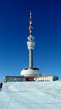 A large telecommunications tower with multiple antennas stands prominently against a clear, deep blue sky. The base of the tower has a circular structure and is surrounded by a snow-covered landscape. A few people are walking towards the building.