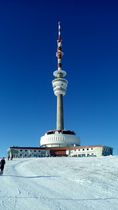 A large telecommunications tower with multiple antennas stands prominently against a clear, deep blue sky. The base of the tower has a circular structure and is surrounded by a snow-covered landscape. A few people are walking towards the building.
