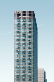 A tall, modern skyscraper with a grid-like facade dominates the image. The upper part of the building displays the names 'RTE' and 'Tarkett'. A clear blue sky provides a backdrop, and a faint contrail is visible. Another high-rise building is partially visible in the background.
