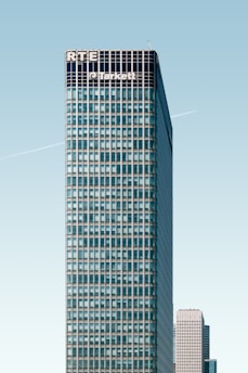 A tall, modern skyscraper with a grid-like facade dominates the image. The upper part of the building displays the names 'RTE' and 'Tarkett'. A clear blue sky provides a backdrop, and a faint contrail is visible. Another high-rise building is partially visible in the background.