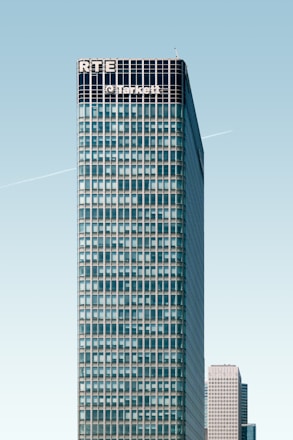 A tall, modern skyscraper with a grid-like facade dominates the image. The upper part of the building displays the names 'RTE' and 'Tarkett'. A clear blue sky provides a backdrop, and a faint contrail is visible. Another high-rise building is partially visible in the background.