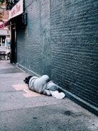 A person is lying on the sidewalk against a brick wall, covered in a grey hoodie. They are lying on cardboard and have a white bag nearby. The scene is urban, with storefronts visible in the background.