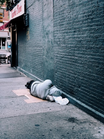 A person is lying on the sidewalk against a brick wall, covered in a grey hoodie. They are lying on cardboard and have a white bag nearby. The scene is urban, with storefronts visible in the background.