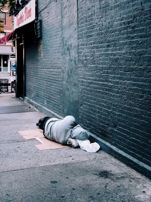 A person is lying on the sidewalk against a brick wall, covered in a grey hoodie. They are lying on cardboard and have a white bag nearby. The scene is urban, with storefronts visible in the background.