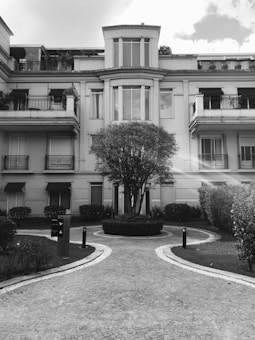 A monochromatic image of a residential building with a symmetrical facade. The architecture features multiple balconies with ornate railings. A manicured garden with a paved pathway leading to the entrance is in the foreground. A large, well-trimmed tree is centered in front of the building.