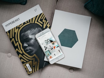 A magazine titled 'HYPEBEAST' with bold, abstract patterns featuring a close-up portrait lies on a couch beside an iPhone displaying a grid of application icons. A minimalistic book with a hexagonal design on the cover is also present.