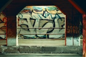 A concrete section of a building is covered with vibrant graffiti art, featuring bold letters and abstract shapes. The graffiti includes various colors and styles, giving a sense of urban culture. Metal railings are positioned in front of the graffiti, suggesting an industrial setting.