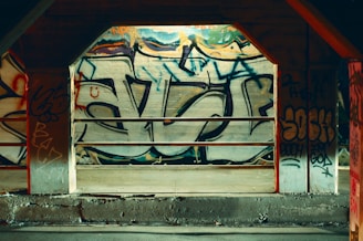 A concrete section of a building is covered with vibrant graffiti art, featuring bold letters and abstract shapes. The graffiti includes various colors and styles, giving a sense of urban culture. Metal railings are positioned in front of the graffiti, suggesting an industrial setting.