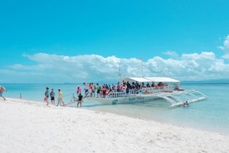 A group of people is boarding a large white outrigger boat on a pristine sandy beach. The turquoise ocean is calm, stretching out to a horizon filled with fluffy white clouds under a bright blue sky.