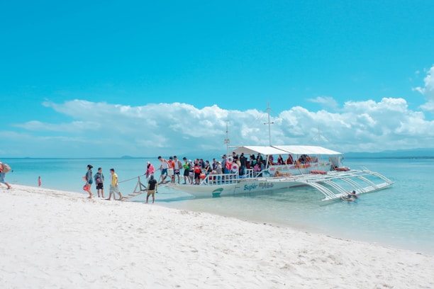 A group of people is boarding a large white outrigger boat on a pristine sandy beach. The turquoise ocean is calm, stretching out to a horizon filled with fluffy white clouds under a bright blue sky.