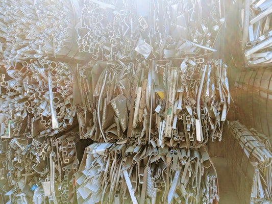 Stacks of various metal profiles and extrusions, including pipes and rectangular tubes, arranged vertically in a warehouse setting. The surfaces appear reflective and are bathed in soft, diffused light.