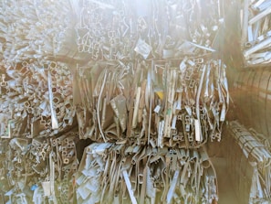 Stacks of various metal profiles and extrusions, including pipes and rectangular tubes, arranged vertically in a warehouse setting. The surfaces appear reflective and are bathed in soft, diffused light.