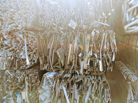 Stacks of various metal profiles and extrusions, including pipes and rectangular tubes, arranged vertically in a warehouse setting. The surfaces appear reflective and are bathed in soft, diffused light.