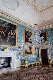 A lavishly decorated room featuring ornate blue damask wallpaper, several large classical paintings in gilded frames, an intricate crystal chandelier, and opulent gold-trimmed furniture. The ceiling is adorned with detailed plasterwork and a carpet with intricate patterns covers the floor. A grand fireplace with sculptural elements and a clock is positioned against the wall.