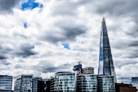 A modern cityscape featuring a prominent skyscraper with a sharp, pointed design, surrounded by several other glass-clad buildings under a predominantly cloudy sky with patches of blue visible.