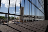 A metal fence in the foreground with vertical bars frames a construction site, where various materials and machinery are visible. In the background, an industrial building with a tower stands under a partly cloudy blue sky, surrounded by greenery and other urban structures.
