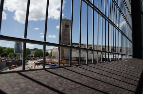 A metal fence in the foreground with vertical bars frames a construction site, where various materials and machinery are visible. In the background, an industrial building with a tower stands under a partly cloudy blue sky, surrounded by greenery and other urban structures.