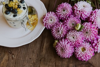 A glass jar filled with creamy yogurt topped with blueberries and a drizzle of honey sits on a white plate. Next to it is a cluster of vibrant pink chrysanthemums placed on a wooden surface.