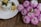A glass jar filled with creamy yogurt topped with blueberries and a drizzle of honey sits on a white plate. Next to it is a cluster of vibrant pink chrysanthemums placed on a wooden surface.