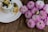 A glass jar filled with creamy yogurt topped with blueberries and a drizzle of honey sits on a white plate. Next to it is a cluster of vibrant pink chrysanthemums placed on a wooden surface.