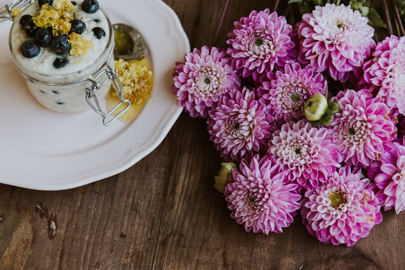 A glass jar filled with creamy yogurt topped with blueberries and a drizzle of honey sits on a white plate. Next to it is a cluster of vibrant pink chrysanthemums placed on a wooden surface.