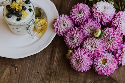 A glass jar filled with creamy yogurt topped with blueberries and a drizzle of honey sits on a white plate. Next to it is a cluster of vibrant pink chrysanthemums placed on a wooden surface.