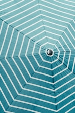 A close-up view of the underside of a large umbrella, featuring a turquoise color with white, evenly spaced radial stripes extending from the center. The fabric appears taut, with the frame of the umbrella faintly visible, creating a geometric pattern.