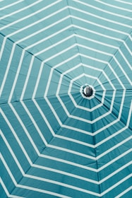 A close-up view of the underside of a large umbrella, featuring a turquoise color with white, evenly spaced radial stripes extending from the center. The fabric appears taut, with the frame of the umbrella faintly visible, creating a geometric pattern.