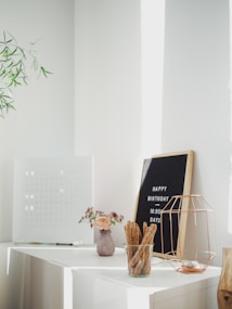 A bright and minimalist interior space with a modern aesthetic. On a white surface, there is a small pink vase containing a bouquet of flowers, a clear jar filled with sesame-covered breadsticks, and a wireframe candle holder. A framed letter board displays a 'HAPPY BIRTHDAY' message and the number of days. A decorative plant branch is visible on the left.