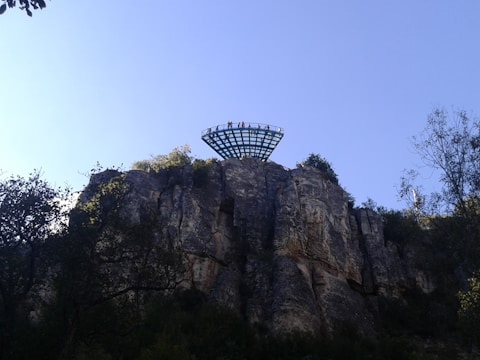 A platform extends over a cliff with several people standing on it, surrounded by trees and vegetation. The rock face is rugged and the sky is clear with a light blue hue.