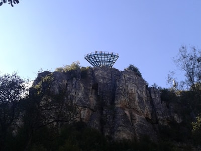 A platform extends over a cliff with several people standing on it, surrounded by trees and vegetation. The rock face is rugged and the sky is clear with a light blue hue.