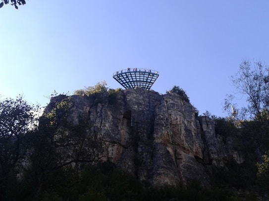 A platform extends over a cliff with several people standing on it, surrounded by trees and vegetation. The rock face is rugged and the sky is clear with a light blue hue.
