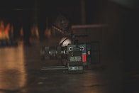 A professional digital camera placed on a dark surface with a lens and other camera equipment visible. The lighting is dim, creating a moody atmosphere with soft reflections on the floor.