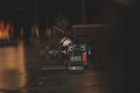 A professional digital camera placed on a dark surface with a lens and other camera equipment visible. The lighting is dim, creating a moody atmosphere with soft reflections on the floor.