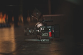 A professional digital camera placed on a dark surface with a lens and other camera equipment visible. The lighting is dim, creating a moody atmosphere with soft reflections on the floor.