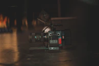 A professional digital camera placed on a dark surface with a lens and other camera equipment visible. The lighting is dim, creating a moody atmosphere with soft reflections on the floor.