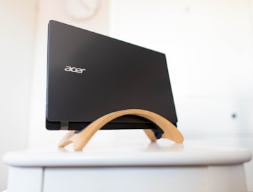 A black laptop with the logo 'acer' is placed in a wooden stand on a white surface. The background is softly blurred, providing a clean and minimalist setting.