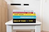 A stack of five books is neatly arranged on a white table. The books are assorted in color and size, featuring prominent titles and authors visible on their spines. The titles include 'Outliers,' 'Crucial Conversations,' 'Deep Work,' 'Getting Things Done,' and 'Tools of Titans.' The background is softly blurred, drawing focus to the books.