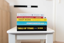 A stack of five books is neatly arranged on a white table. The books are assorted in color and size, featuring prominent titles and authors visible on their spines. The titles include 'Outliers,' 'Crucial Conversations,' 'Deep Work,' 'Getting Things Done,' and 'Tools of Titans.' The background is softly blurred, drawing focus to the books.