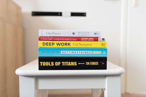 A stack of five books is neatly arranged on a white table. The books are assorted in color and size, featuring prominent titles and authors visible on their spines. The titles include 'Outliers,' 'Crucial Conversations,' 'Deep Work,' 'Getting Things Done,' and 'Tools of Titans.' The background is softly blurred, drawing focus to the books.