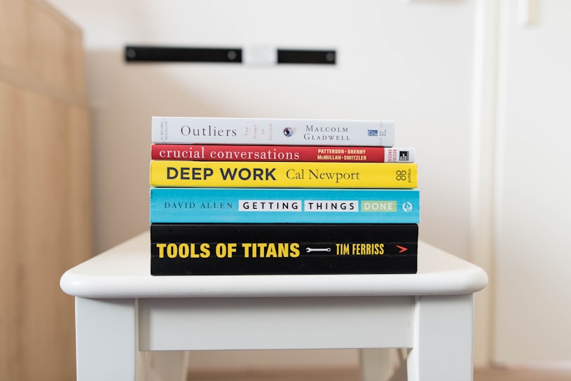 A stack of five books is neatly arranged on a white table. The books are assorted in color and size, featuring prominent titles and authors visible on their spines. The titles include 'Outliers,' 'Crucial Conversations,' 'Deep Work,' 'Getting Things Done,' and 'Tools of Titans.' The background is softly blurred, drawing focus to the books.