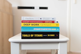 A stack of five books is neatly arranged on a white table. The books are assorted in color and size, featuring prominent titles and authors visible on their spines. The titles include 'Outliers,' 'Crucial Conversations,' 'Deep Work,' 'Getting Things Done,' and 'Tools of Titans.' The background is softly blurred, drawing focus to the books.