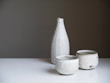 A set of minimalist ceramic tableware, consisting of a tall bottle and two cups, displayed on a white surface against a neutral gradient background. The ceramic pieces are speckled with subtle dark spots and have a rough, rustic texture.