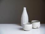 A set of minimalist ceramic tableware, consisting of a tall bottle and two cups, displayed on a white surface against a neutral gradient background. The ceramic pieces are speckled with subtle dark spots and have a rough, rustic texture.