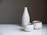 A set of minimalist ceramic tableware, consisting of a tall bottle and two cups, displayed on a white surface against a neutral gradient background. The ceramic pieces are speckled with subtle dark spots and have a rough, rustic texture.