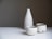 A set of minimalist ceramic tableware, consisting of a tall bottle and two cups, displayed on a white surface against a neutral gradient background. The ceramic pieces are speckled with subtle dark spots and have a rough, rustic texture.