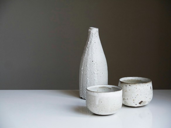 A set of minimalist ceramic tableware, consisting of a tall bottle and two cups, displayed on a white surface against a neutral gradient background. The ceramic pieces are speckled with subtle dark spots and have a rough, rustic texture.