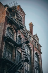 A red brick building with large arched windows and a black metal fire escape attached to the exterior. The architectural style is reminiscent of early 20th-century industrial design, with intricate window framing and details that add to its historic character.