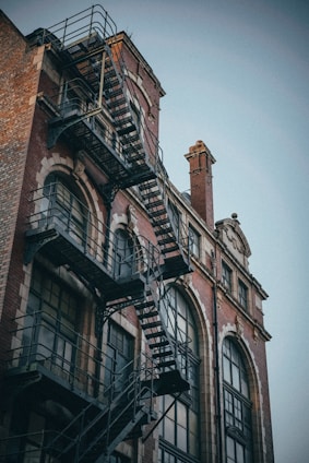 A red brick building with large arched windows and a black metal fire escape attached to the exterior. The architectural style is reminiscent of early 20th-century industrial design, with intricate window framing and details that add to its historic character.
